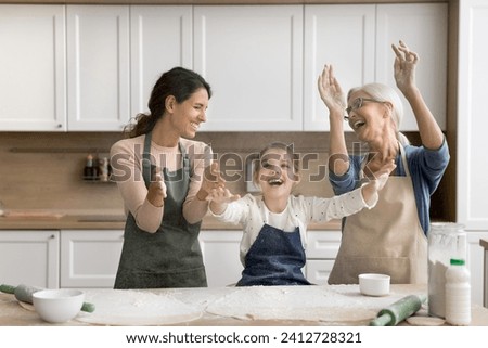 Cheerful excited kid girl, mom and grandma throwing flour powder over baking table, clapping hands, making floury cloud, laughing, having fun, enjoying family leisure, playtime, cooking hobby Royalty-Free Stock Photo #2412728321