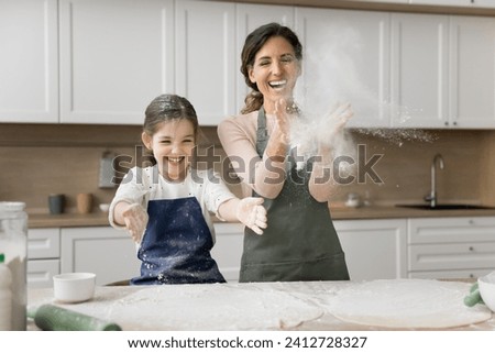 Excited mom and cheerful little daughter girl in aprons throwing flour while baking in home kitchen, having fun, laughing, clapping floury hands, enjoying culinary hobby, childhood, motherhood Royalty-Free Stock Photo #2412728327
