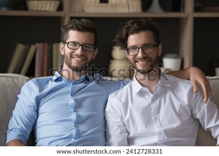 Young adult twins brothers, hugging, sit together on couch in living room, smiling, looking at camera, enjoy friendly communication, showing unity, keeping good close relationships. Family ties, bond