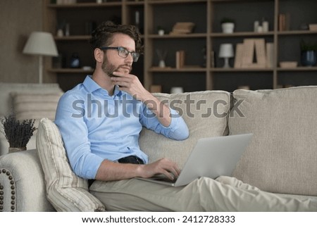 Serious thoughtful 35s man sit on sofa with wireless computer, get message, thinks on answer or creative online task, learn new AI program, looks pensive. Blogging, telework using laptop, modern tech Royalty-Free Stock Photo #2412728333