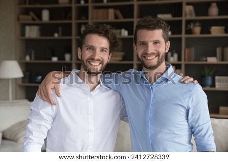 Portrait of two millennial twin siblings brothers in casual shirts standing in living room, hugging smile staring at camera. Family ties, good, friendly and harmonic relationships, support and love
