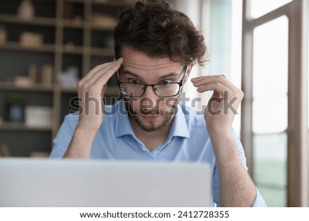 Close up face of young man in glasses stare wide-eyed at laptop screen, feel shocked by received news, confused or surprised by unexpected error, broken computer, overwhelmed with heap of scam message Royalty-Free Stock Photo #2412728355