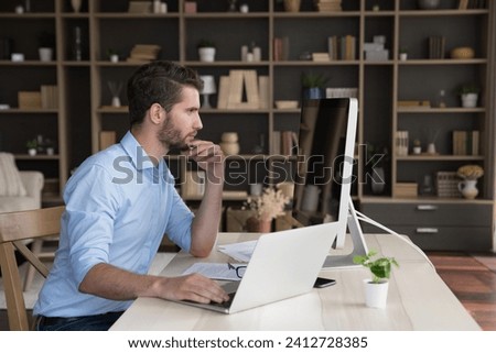 Thoughtful millennial 35s man working sit at desk looks at device screen, learn AI app, artificial intelligence program working in cozy homeoffice, review sales statistics, doing SEO optimization job