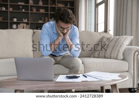 Stressed single man feels frustrated due to overspend, high household rates, unpaid taxes, lack of finances to pay domestic bills sit at table with laptop and heap of papers. Penalty, eviction, crisis Royalty-Free Stock Photo #2412728389