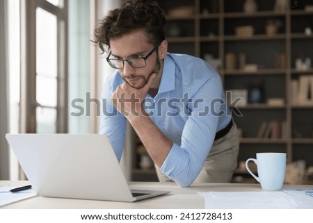 Pensive serious businessman looks at laptop screen, read e-mail, check correspondence answer to client by e-mail, working in modern home-office, review sales results, prepare task. Telework, workflow Royalty-Free Stock Photo #2412728413