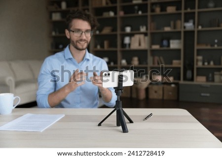Young man in glasses sit at desk, makes speech, looking at smartphone camera, take part in online stream, record webinar, share information. Social media icon, influencing, vlogging, videocall event Royalty-Free Stock Photo #2412728419