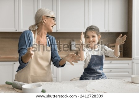 Excited grandmother and happy little granddaughter girl in aprons throwing flour over table with ingredients laughing, having fun, clapping floury hands for making cloud, playing while cooking Royalty-Free Stock Photo #2412728509