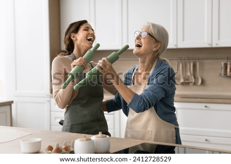 Joyful older mom and excited adult daughter having fun in kitchen, acting singers while baking pastry, singing song at roller mics, shouting, laughing, having fun, enjoying family cooking, friendship Royalty-Free Stock Photo #2412728529