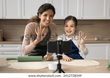 Family cooking blogger mom and happy cute kid girl in aprons baking in home kitchen together, recording video for blog on smartphone, waving hands hello at camera, smiling, laughing Royalty-Free Stock Photo #2412728549