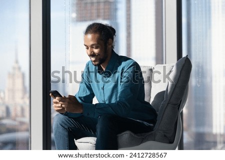 Things are going well. Smiling mixed race businessman sit at comfy office chair by picture window use social media app on cell. Happy young black male employee networking with colleague at online chat