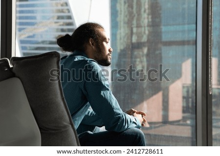 Creating idea. Pensive biracial businessman sit in cozy ergonomic armchair by window of corporate office look at distance. Young black guy team leader think on business vision strategy plan. Copyspace
