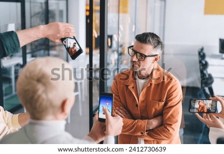 Confident male entrepreneur in optical eyewear for vision correction posing during paparazzi photo session in office workspace, unrecognizable people with smartphones shooting video of corporate boss