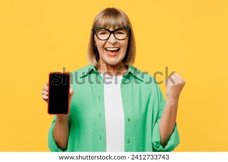 Elderly blonde woman 50s years old wear green shirt glasses casual clothes hold use mobile cell phone with blank screen area do winner gesture isolated on plain yellow background. Lifestyle concept