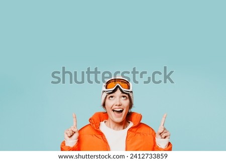 Skier young woman wear warm padded windbreaker jacket hat ski goggles mask point index finger overhead on area travel rest spend weekend winter season in mountains isolated on plain blue background