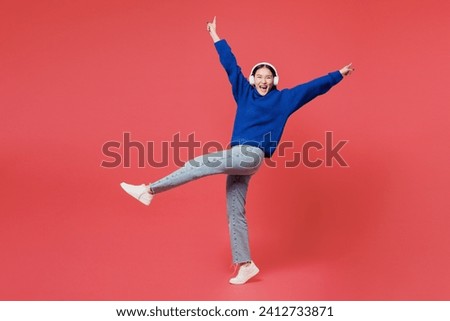 Full body cool young woman of Asian ethnicity she wear blue sweater casual clothes listen to music in headphones raise up leg spread hands isolated on plain pastel pink background. Lifestyle concept