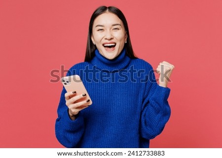 Young smiling happy woman of Asian ethnicity she wear blue sweater casual clothes hold in hand use mobile cell phone do winner gesture isolated on plain pastel light pink background. Lifestyle concept