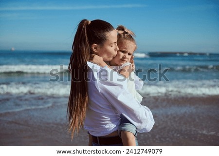 Authentic portrait of a beautiful young Caucasian woman carrying, kissing and hugging her little daughter, enjoying happy time together while walking on the beach. People. Lifestyle. Leisure activity