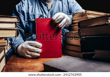 Photo of antiquarian man in a blue shirt and white protective gloves holding a red book and sitting by a table with books pile. Royalty-Free Stock Photo #2412763905