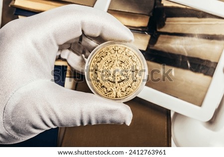 Photo of a person's male hand in white gloves holding a golden antique aztec or mayan coin on stack of books and magnifyng glass background. Numismatics hobby concept.