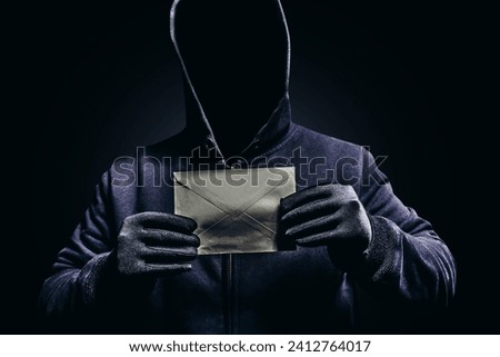 Photo of scary shaded hooded man holding paper letter on dark background. Royalty-Free Stock Photo #2412764017