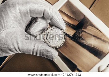 Photo of a person's male hand in white gloves holding a soviet 1970 ruble coin on stack of books and magnifyng glass background. Numismatics hobby concept. Royalty-Free Stock Photo #2412764213