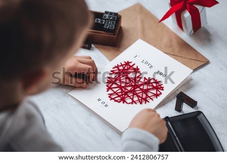 Little kid preparing diy handmade cute post card for Mother's Day with a message I love you mom. Red hear embroidering, printing. Concept of simple presents, family values, hobby, leisure activity Royalty-Free Stock Photo #2412806757