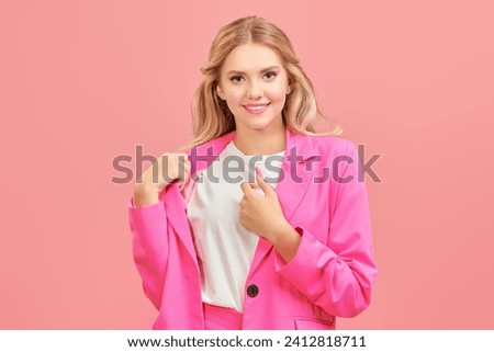A pretty young woman with wavy blonde hair and gentle pink makeup, dressed in a pink suit, smiles charmingly. Pink studio background with copy space.