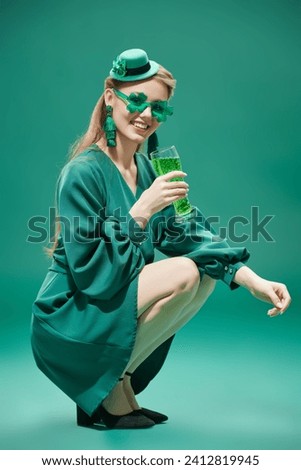 Party for St. Patrick's Day. A cheerful girl, dressed in a green dress, a hat and green clover-shaped glasses, holds a glass of beer. Full-length studio portrait against a green background.