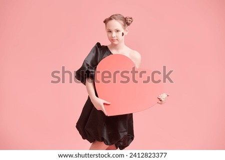 A cute teenage girl in an elegant black dress posing emotionally on a pink background with a red heart in her hands. Valentine's Day. Copy space.