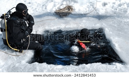 Two men equipped for winter under ice diving are swimming in an ice hole, tightened with a safety rope, ready to immerse, bubbles coming up from water Royalty-Free Stock Photo #2412841453