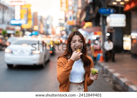 Young Asian tourists standing and eating Thai desserts. Young beautiful tourists in Chinatown street food market, Bangkok, Thailand Royalty-Free Stock Photo #2412849401