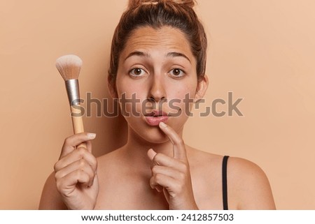 Photo of surprised European woman holds cosmetic brush going to apply powder on face feels amazed stands bare shouldered isolated over brown background. Daily makeup and cosmetology concept.