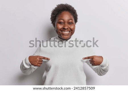 People and positive emotions. Indoor portrait of young happy smiling African american woman standing in centre isolated on white background wearing hoodie pointing at bosom as space for inscription Royalty-Free Stock Photo #2412857509