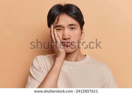 Portrait of displeased Japanese man has dull day keeps hand on cheek showing lack of interest feels bored dressed in casual t shirt isolated over brown background. Negative human emotions concept