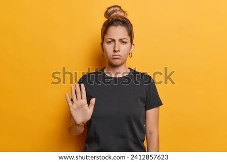 Waist up shot of serious European woman with hair bun makes stop gesture shows denial sign being dissatisfied with something wears black t shirt isolated over yellow background. Stop abusing me