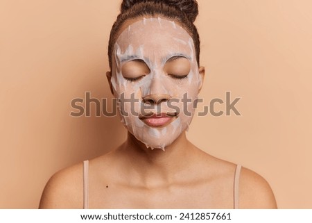 Studio shot of young beautiful calm relaxed European girl with closed eyes wearing undershirt keeping hair in bun doing beauty procedure applying face mask standing isolated on beige background