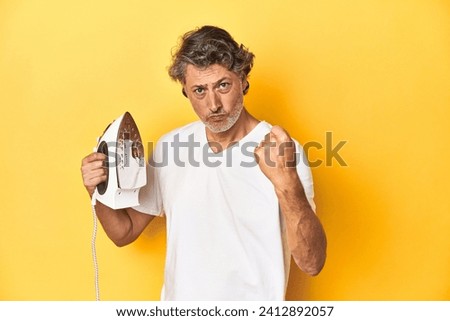 Man with iron in hand on a yellow background showing fist to camera, aggressive facial expression.