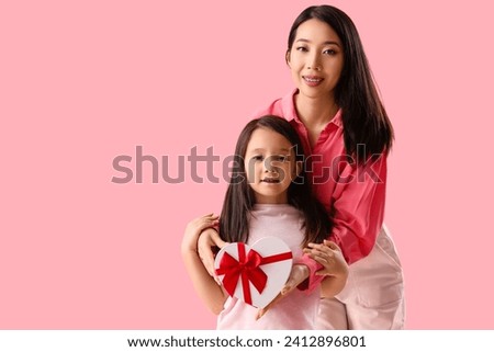 Young Asian woman and her little daughter with gift for Valentine's day on pink background
