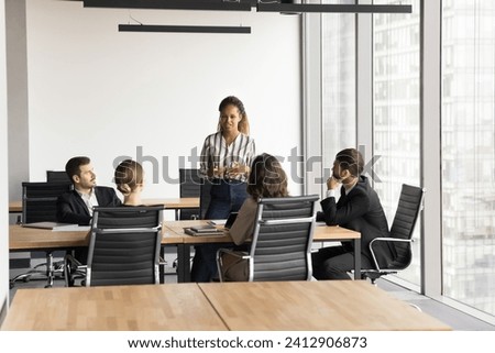 Positive motivated young Black business professional holding meeting with team of colleagues, partners, standing at large table in modern office space, speaking, presenting ideas for brainstorming Royalty-Free Stock Photo #2412906873