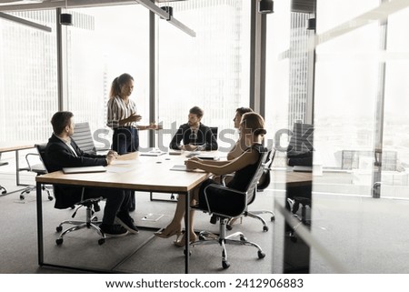 Wide shot of diverse millennial business team meeting at large table in contemporary city office interior. African project manager offering ideas, teamwork plan, talking to colleagues Royalty-Free Stock Photo #2412906883