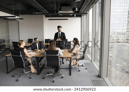 Serious business leader man in formal suit speaking to team on meeting in modern urban office space, standing at table, offering cooperation plan to diverse colleagues. Full length wide shot Royalty-Free Stock Photo #2412906923