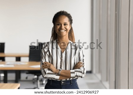 Positive beautiful young Black business woman posing in office with hands folded, looking at camera with toothy smile. Happy female entrepreneur, professional, worker girl head shot portrait