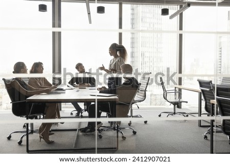 Diverse business group meeting at table in modern urban office, listening to young female African American leader, discussing work project, planning cooperation, teamwork. Through glass view Royalty-Free Stock Photo #2412907021