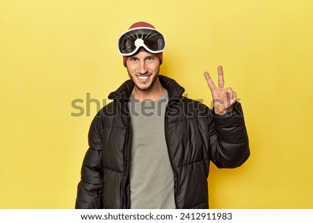 Man in winter coat with snow goggles on yellow showing number two with fingers.
