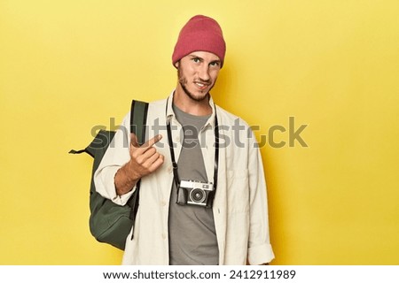 Man with vintage camera and backpack on yellow pointing with finger at you as if inviting come closer.