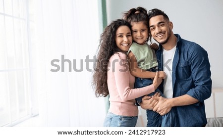 Portraif Of Happy Arabic Parents Posing With Their Little Daughter At Home, Loving Middle Eastern Family Of Three Embracing Near Window And Smiling At Camera, Enjoying Time Together, Copy Space Royalty-Free Stock Photo #2412917923