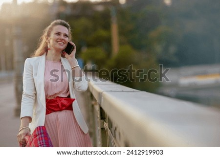 happy modern woman in pink dress and white jacket in the city speaking on a smartphone on the bridge. Royalty-Free Stock Photo #2412919903