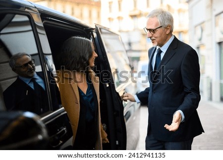Senior businessman is engaging in a friendly conversation with a female assistant while entering a luxurious limousine Royalty-Free Stock Photo #2412941135