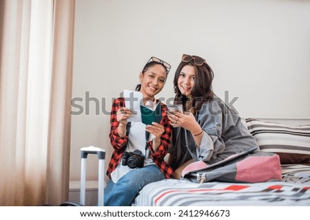 two smiling women holding tickets and cell phones while resting on a hotel room bed