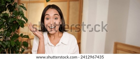 Portrait of asian girl looking surprised, holding mobile phone with amazed face, great news on smartphone, sitting in cafe with book.